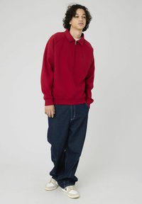 Pull en polaire rouge avec une demi-fermeture éclair, présentant un col montant et des manches longues. Associé à un jean en denim foncé ample et des baskets blanches.