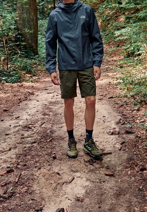 Persona con chaqueta azul marino, pantalones cortos color oliva y zapatillas de senderismo de pie en un camino de tierra en un bosque denso.