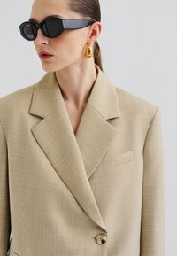 Beige plaid blazer with a notched collar, single button closure, and a pocket. Paired with black angular sunglasses and gold hoop earrings.