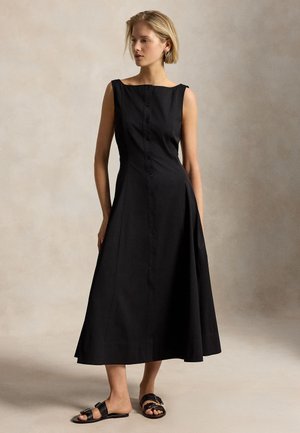 Žena oblečená v čiernych midi šatách bez rukávov s rozšírenou sukňou a čiernymi nízkymi sandálmi, stojaca na neutrálnom pozadí.