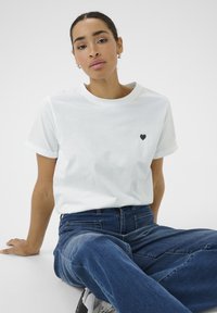 Camiseta de algodón blanca con mangas enrolladas, que presenta un pequeño logo de corazón negro en el pecho. Se lleva con jeans de mezclilla azules.