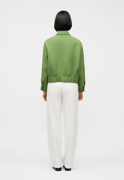 Personne aux cheveux courts noirs vue de dos portant une veste verte et un pantalon blanc à jambes larges sur un fond uni.