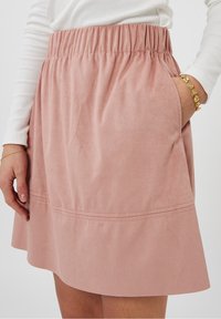 Jupe en daim rose avec une taille élastique, de forme évasée et poches sur les côtés. Elle présente une texture lisse et des coutures discrètes à l'ourlet.