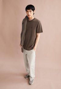 Braunes Baumwoll-T-Shirt mit einem kleinen schwarzen Band-Logo, kombiniert mit hellgrauen Cargo-Hosen und grauen Sneakers. Das Modell steht vor einem neutralen Hintergrund.