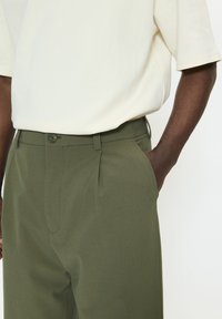 Olívazöld színű, testhezálló szabású nadrág, egyetlen elöl hajtással és oldalzsebekkel, krémszínű rövid ujjú inggel párosítva.