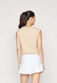 Beige mouwloos topje met een V-hals en een wit logo op de achterkant, gecombineerd met een witte flared rok. Soepele stoftextuur.