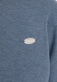 Tissu texturé bleu avec une petite étiquette ovale en métal argenté cousue, gravée du mot « Smilodox » en cursive.