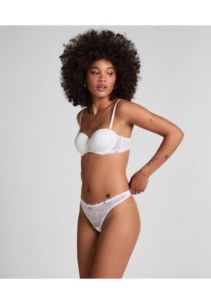 Frau richtet weißen Spitzen-String und passenden trägerlosen BH, stehend vor schlichtem hellgrauem Hintergrund.