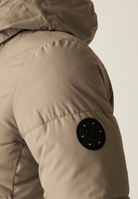Beige puffer takki, jossa on huppu ja tikattu pinta. Vasen hiha sisältää pyöreän mustan logoläpatchi, jossa on valkoisia yksityiskohtia.