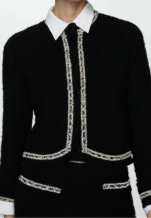 Conjunto de chaqueta y pantalones negros con textura y ribete de tweed blanco, usado sobre una camisa blanca con cuello abotonada al frente.