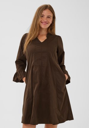 Femme souriante, mains dans les poches, portant une robe marron à manches longues, longueur genou, avec des poignets volantés et un col en V.