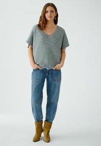 Grauer Strickpullover mit V-Ausschnitt und kurzen Ärmeln, kombiniert mit blauen Jeans und senfgelben Stiefeletten. Leichte strukturierte Überlagerung am Saum.