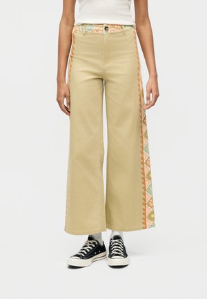 Γυναίκα που φοράει μπεζ παντελόνι με φαρδύ πόδι και γεωμετρικά διακοσμητικές πλαϊνές λεπτομέρειες, λευκές κάλτσες, μαύρα αθλητικά παπούτσια και λευκό αμάνικο τοπ.