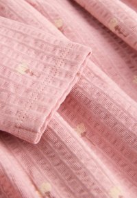 Světle růžová textilie s texturovaným povrchem, zdobená malými zlatými srdíčkovými vzory. Viditelné složené okraje a švy.