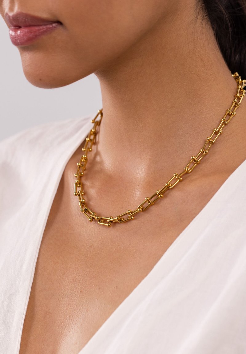 Gros plan d'une personne portant un collier chaîne en or avec de petits accents en forme de boule sur un haut blanc à col en V.