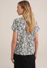 Blusa a maniche corte in verde oliva con un motivo floreale bianco e lavanda. Tessuto leggero con una vestibilità morbida e orlo arrotondato.