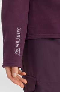 Lähedalt vaadatuna on käsi, millel on seljas lilla fliisist varrukas, mille mansetil on brodeeritud "POLARTEC". Käsi on koos sobivate lilla pükstega, millel on nööbiga tasku.