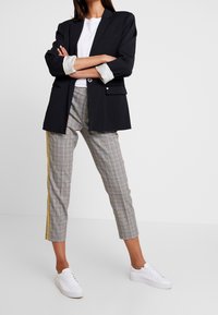 Czarna marynarka z pojedynczym guzikiem, zestawiona z szarymi spodniami w kratkę z żółtymi paskami po bokach i białymi trampkami. Prosty, dopasowany krój.