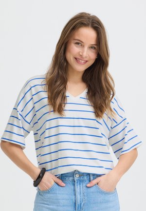 Femme souriante aux longs cheveux bruns portant un t-shirt blanc à col en V avec des rayures bleues et un jean bleu clair, mains dans les poches, devant un fond uni.