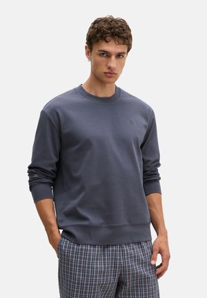Giovane uomo con capelli ricci che indossa una felpa blu scuro a maniche lunghe e pantaloni a quadri grigi, in piedi con una mano in tasca.