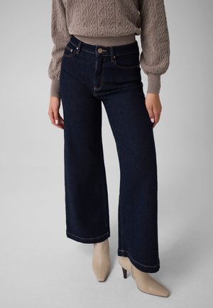 Femme portant un jean large foncé, des bottines beiges et un pull taupe en maille torsadée avec manches ballon.
