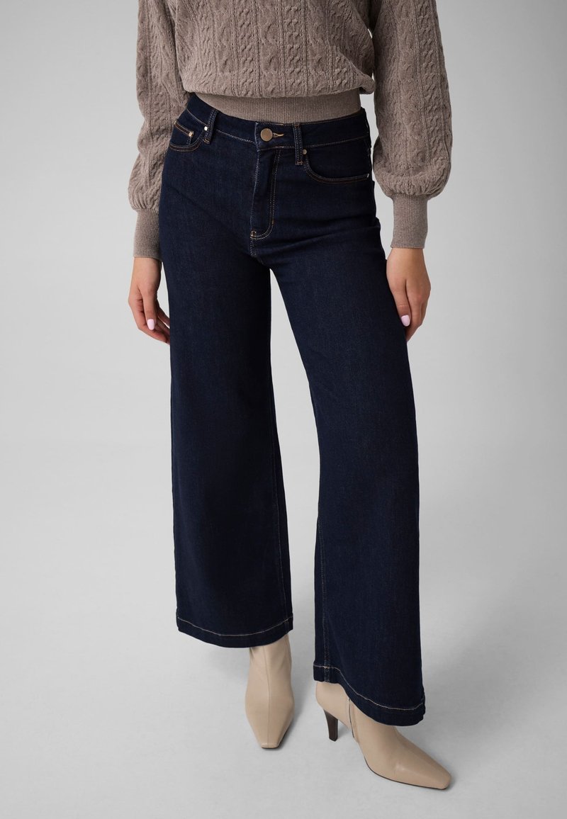 Femme portant un jean large foncé, des bottines beiges et un pull taupe en maille torsadée avec manches ballon.