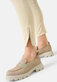 Zapatos tipo mocasín beige con suela gruesa, que presentan una textura suave y una correa decorativa. Pantalones beige con detalle de cremallera en el tobillo.