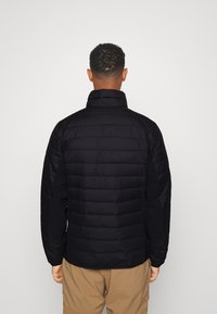 Svart vadderad jacka med hög krage. Designen har horisontella quiltade sektioner och elastiska ärmar. Bärs över beiga byxor.