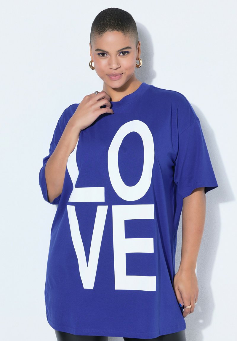 Persona con cabello corto que lleva pendientes grandes de aro y una camisa azul holgada con la palabra "LOVE" en letras blancas.