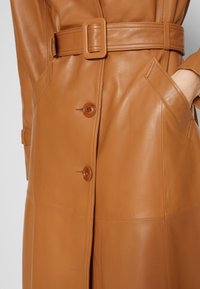 Manteau trench en cuir marron avec ceinture, doté de boutons et de poches latérales. La texture est lisse avec un léger éclat.