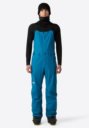 Blue waterproof overalls with adjustable straps, multiple pockets, and a faux-leather black neck gaiter, worn over a black long-sleeve shirt.
