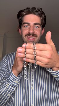 Correntes prateadas seguradas nas mãos, apresentando várias texturas e formas com espigões e elos. Camisa às riscas visível ao fundo.