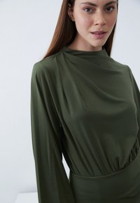 Olivegrünes Kleid mit lockerer Passform, langen Ärmeln, hohem Kragen und geraffter Taille, aus glattem, leichtem Stoff gefertigt.