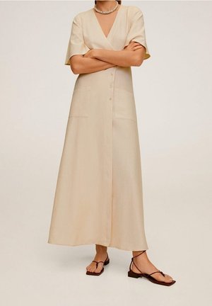 Robe portefeuille beige à manches courtes avec un décolleté en V, des détails à boutons, des poches latérales et une coupe fluide. Portée avec des sandales plates simples.