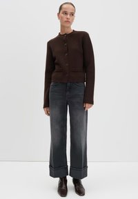 Woman standing wearing a dark brown buttoned cardigan, gray wide-leg cuffed jeans, and dark brown ankle boots on a plain light gray background.