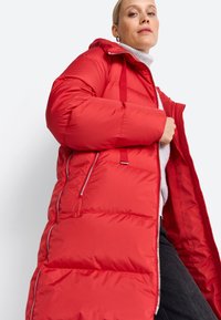 Femeie îmbrăcată cu o jachetă puffer roșie lungă peste un pulover alb cu guler înalt și pantaloni negri, pozând pe un fundal simplu.