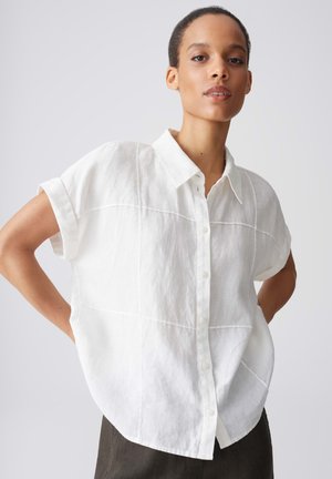 Vrouw met kort haar die een losse witte, kortemouws overhemd met knopen en donkere broek draagt, staat met handen in de zij tegen een effen achtergrond.