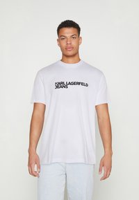 Valge puuvillane T-särk, millel on must trükitud logo "KARL LAGERFELD JEANS", mis on rinnal. Särgil on ümmargune kaelus ja lühikesed varrukad.