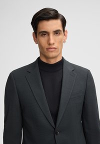 Donkergrijs op maat gemaakt pak met een notch lapel en een enkele knoop, gedragen over een zwart ribbel turtleneck. Glad textuur en gestructureerde pasvorm.