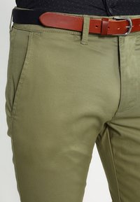 Olive green pants made of smooth fabric with a fitted design. Features a brown leather belt and two front pockets.