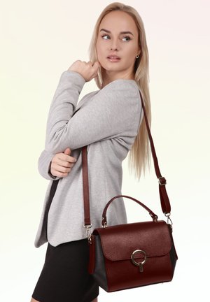 Blond vrouw in een grijze blazer en zwarte rok met een donkerrode leren handtas met schouderriem, kijkt opzij.