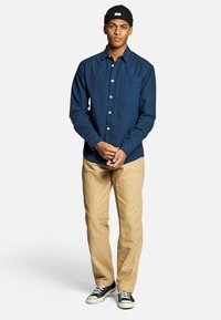 Camicetta blu navy a bottoni con taschino frontale, abbinata a pantaloni khaki e sneaker nere. La camicia ha una vestibilità comoda e una texture liscia.