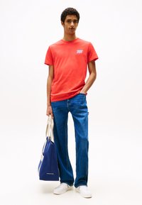 Tricou roșu din bumbac cu logo pe față, asociat cu blugi albaștri. Ținând o geantă tote albastră cu accente albe. Tenisi albi completează look-ul.