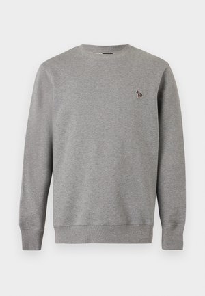 Mann steht mit den Händen in den Taschen, trägt einen grauen Pullover, dunkelblaue Jogginghose und weiße Sneaker vor einem schlichten weißen Hintergrund.