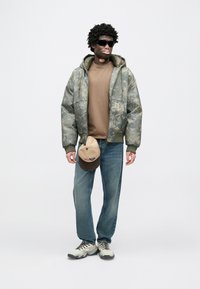 Kurtka puchowa w kamuflażu, brązowa bluza z okrągłym dekoltem, niebieskie dżinsy, beżowa czapka, szaro-czarne trampki. Model stoi pewnie, nosząc okulary przeciwsłoneczne.