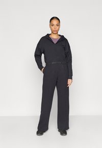 Μαύρο φούτερ με φερμουάρ, cropped σχεδιασμό και ελαστική μέση, σε συνδυασμό με φαρδιά μαύρη παντελόνια. Το μοντέλο ποζάρει μπροστά από απλό φόντο.