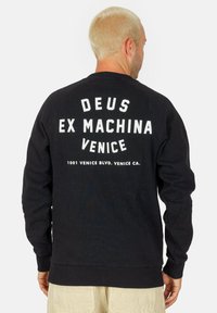 Sudadera negra con texto blanco en la parte trasera: "DEUS EX MACHINA VENICE" y una dirección. Presenta puños y dobladillo acanalados, y está hecha de una tela suave.