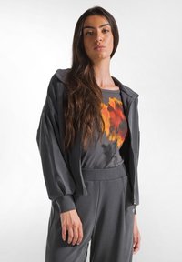 Grijze zip-up jas met brede mouwen, gecombineerd met een donker shirt met een oranje en rode bloemenprint. Gladde stof, relaxte pasvorm.