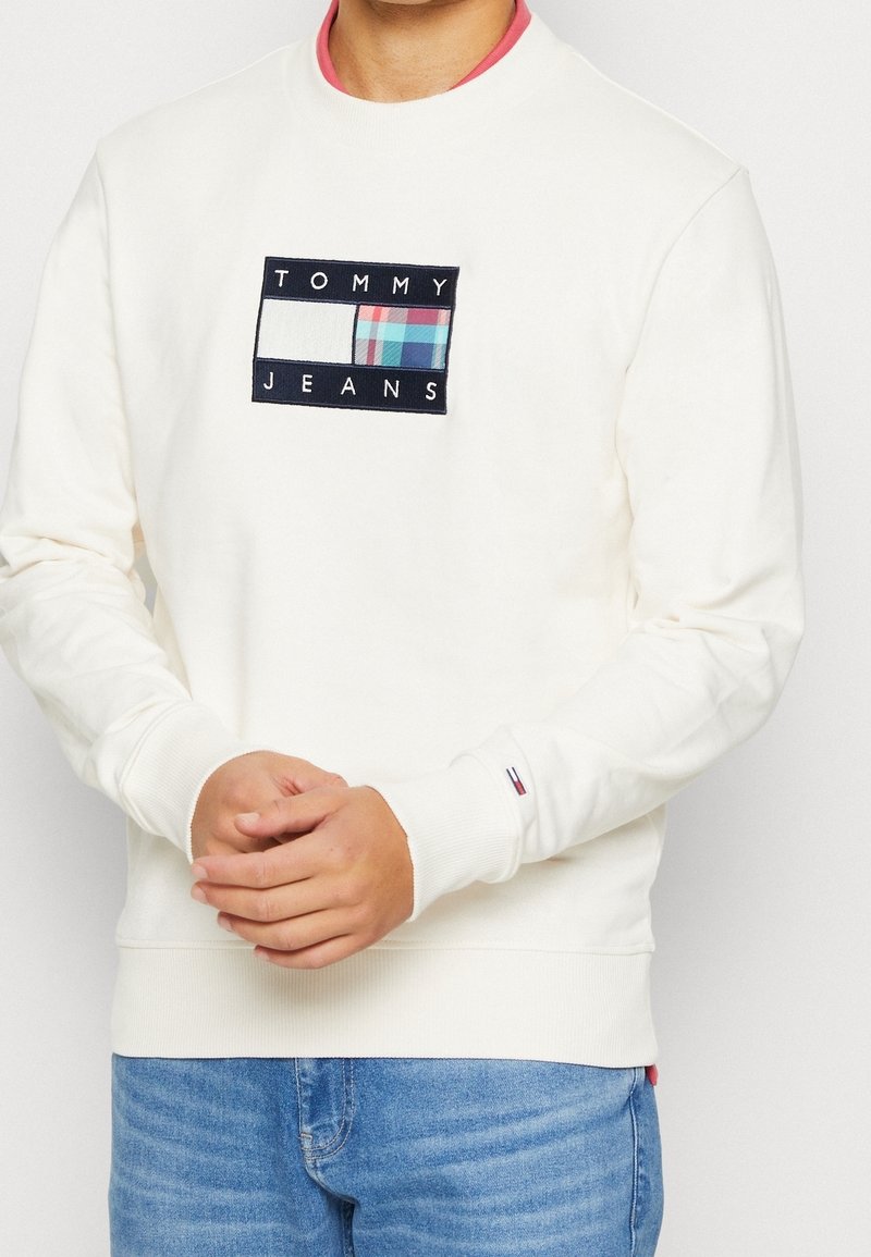 Homme portant un sweat-shirt blanc Tommy Jeans avec un écusson logo coloré sur la poitrine et un jean bleu, les mains jointes sous la taille.