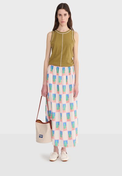 Femme portant un haut sans manches vert olive et une jupe longue à motifs colorés, tenant un sac à main beige et chaussant des baskets blanches.